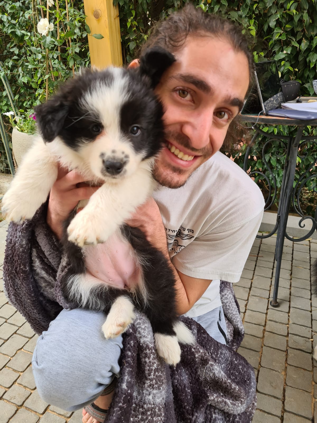 רועי מחזיק גור בורדר קולי שחור־לבן קרוב לפניו ומחייך רחב, בחצר עם צמחייה ירוקה.